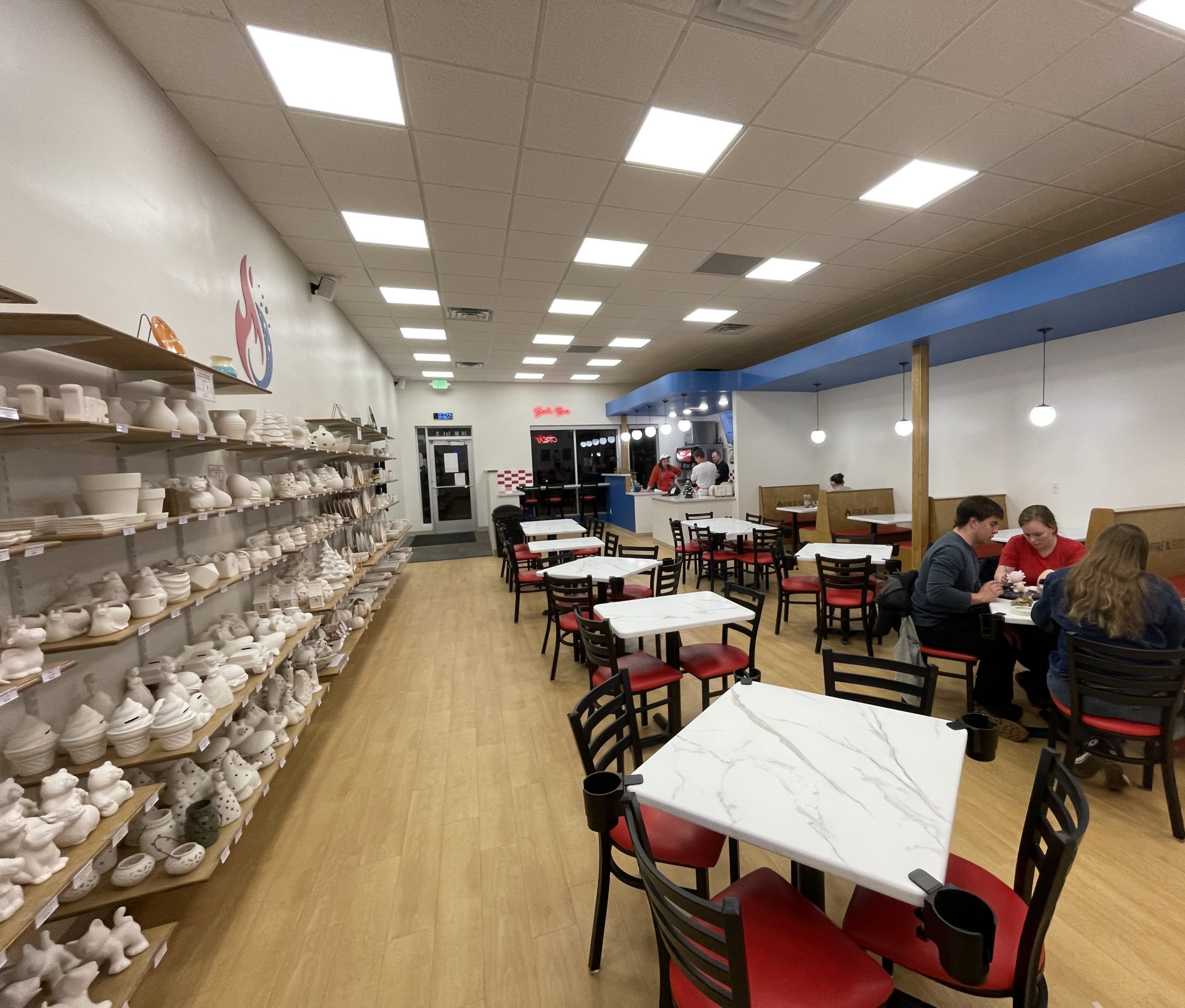 Pottery on shelves on the left and people paiting pottery on the right at tables and pop station in the back.